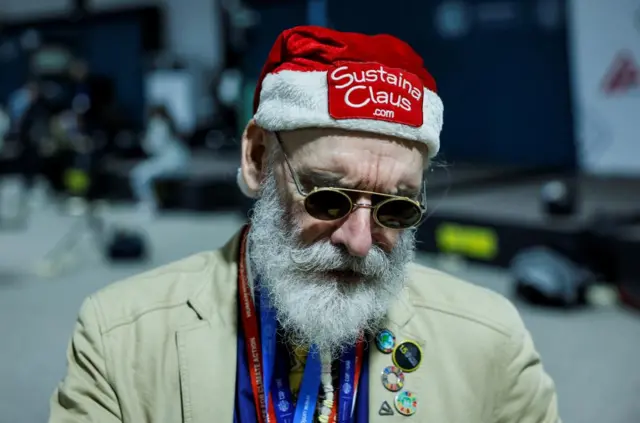 A man wearing a Santa has that says Sustaina Claus on it. He wears sunglasses and badges on his coat