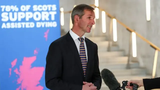 Lib Dem MSP Liam McArthur stands before a poster saying 78% of Scots support assisted dying, he is talking into a TV microphone in the parliament's garden lobby