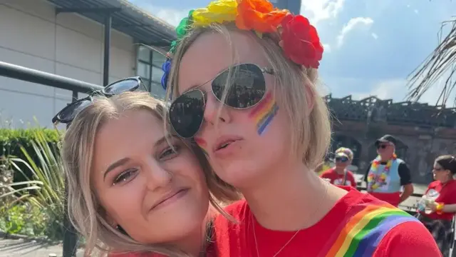 Two young women in bright red t-shirts embrace, smiling, on a sunny day. One wears brightly coloured paper flowers on her head, and has rainbow-coloured stripes painted on her cheeks. They match diagonal stripes printed on her t-shirt. Others dressed in similar fashion stand behind them. It’s a celebratory atmosphere.