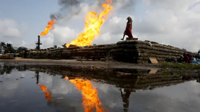 A reflection of two gas flaring furnaces and a woman walking on sand barriers is seen in the pool of oil-smeared water at a flow station in Ughelli, Delta State