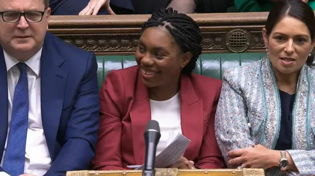 Badenoch smiles as she glances to her right while sitting at PMQs wearing a red blazer, white top and holding some papers