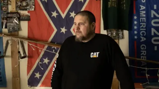 A man with a beard and a black top standing in front of a Confederate flag