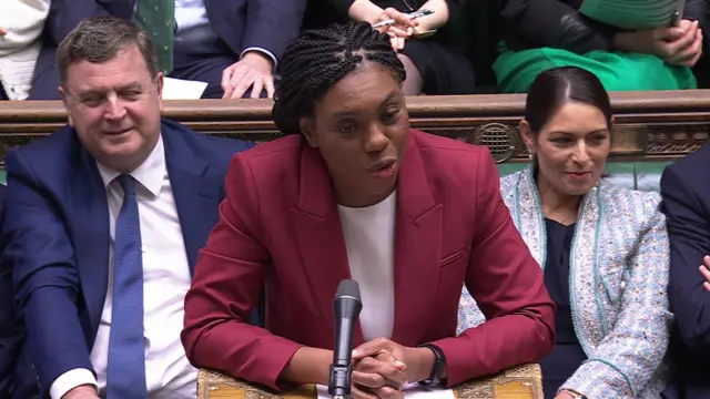 Badenoch wears a red blazer and white top as she leans over the dispatch box with her hands clasped at PMQs