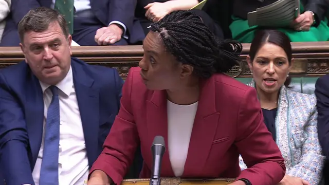 Badenoch wears a red blazer and white top as she leans over the dispatch box and looks to her right