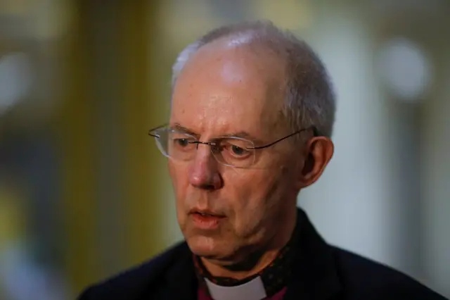 A close-up shot of Welby's face. He is bald and has short grey hair. He is wearing glasses, a clerical collar and a black suit jacket