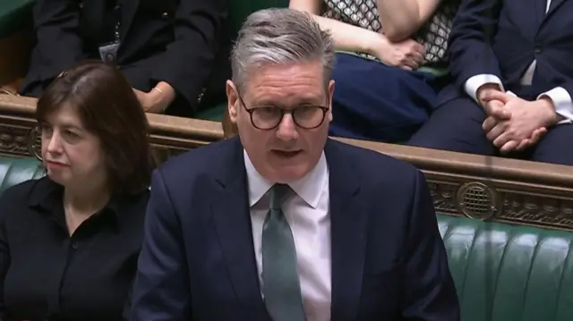 Keir Starmer delivers PMQs in dark suit, white shirt, aquamarine tie. He's wearing glasses and his mouth is partially open as he speaks