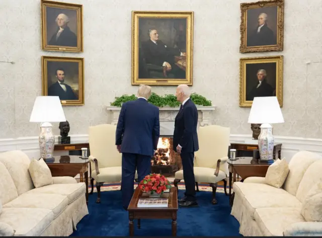 Two men look up at paintings while fire burns in the fireplace behind them