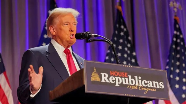 Donald Trump addresses House Republicans from stage. He's standing behind a lectern wearing a dark blue suit, red tie and white shirt. Behind him are four US flags and an illuminated heavy stage curtain