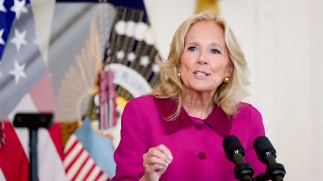 Jill Biden delivers speech behind lectern. She's wearing a hot pink, buttoned up jacket, het right hand slightly raised. Behind her are a US and government flags