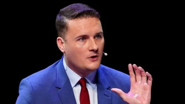 Wes Streeting mid-shot cuts off just below the shoulders. He's in blue jacket, light blue shirt and red tie. His left hand is raised and turned away from him as he speaks, he has a mic attached to his left ear coming down towards the mouth