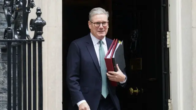 Keir Starmer in dark suit and aquamarine tie holding two folders to his chest, one black to the front and a larger one in red to the back, as he leaves Downing Street