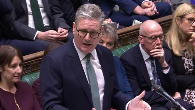 Keir Starmer delivers PMQs in dark suit, white shirt, aquamarine tie. He's wearing glasses and his mouth is partially open as he speaks and gestures with his hands