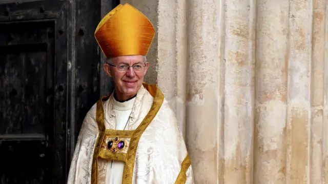 Welby wears a cream coloured cassock with gold detail and an orange bishop's hat. He appears to be standing in a church doorway