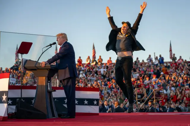 Elon Musk jumps in the air while Donald Trump speaks into a microphone on stage