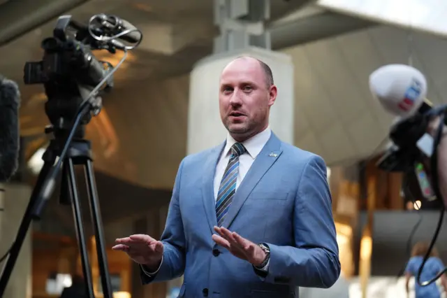 Health Secretary Neil Gray stands behind cameras and microphones in the garden lobby of the Scottish Parliament