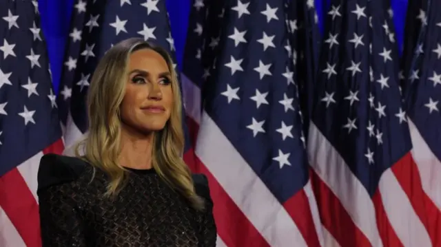 Lara Trump stands to the left of the frame wearing semi-transparent black gown. In background are five US flags