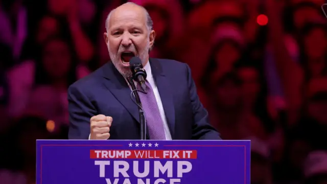 Howard Lutnick, who has grey hair and a short grey beard and is wearing a suit, clenches his fist as he speaks at a Trump rally