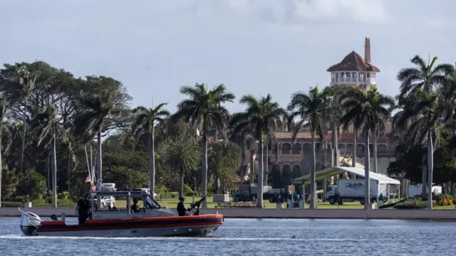 Mar a Lago is seen in the distance. The resort is surrounded by palm trees, and a US Coast Guard boat can be seen patrolling in front of the residence.