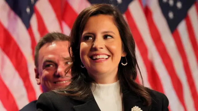 Stefanik on a stage wearing a pearl brooch in front of a background of American flags