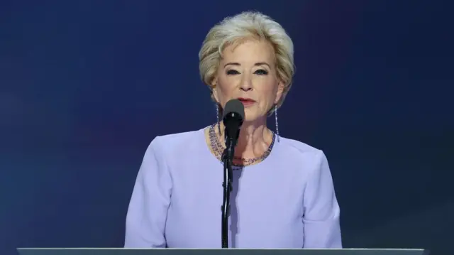 Linda McMahon, who has short blonde hair, wears a lilac long sleeve top and purple beaded jewellery as she speaks on stage