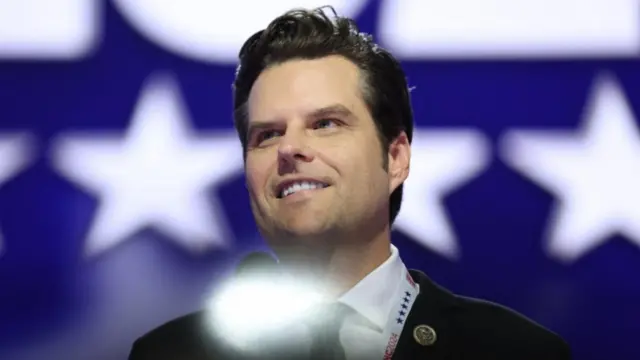 Close up shot (cuts off just below shoulders) of Matt Gaetz on stage smiling. He's wearing a dark suit, white shirt and dark tie partially obscured by a white lens flare at the bottom centre of the image