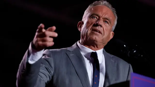 RFK Jr points straight ahead while speaking on stage at a Republican presidential campaign rally. He wears a grey suit, white shirt and navy tie decorated with red flowers.