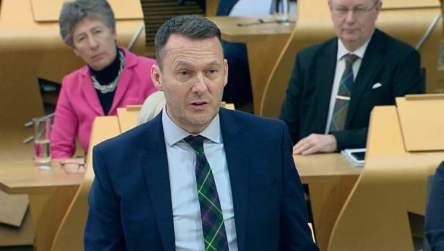 Russell Findlay wearing a dark suit and tartan tie asks a question in the Holyrood chamber