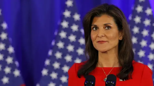 Nikki Haley in red dress stands in front of two mics, three blurred US flags in the background. Mid-shot cuts off just below the shoulders