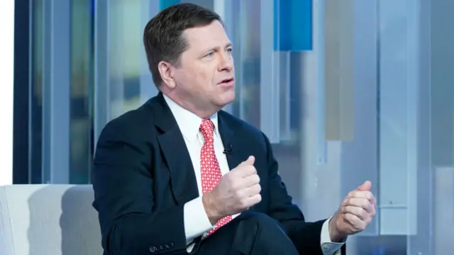 Jay Clayton, with short brown combed hair, wears a black suit with a white shirt and red patterned tie as he sits on a sofa during a TV interview