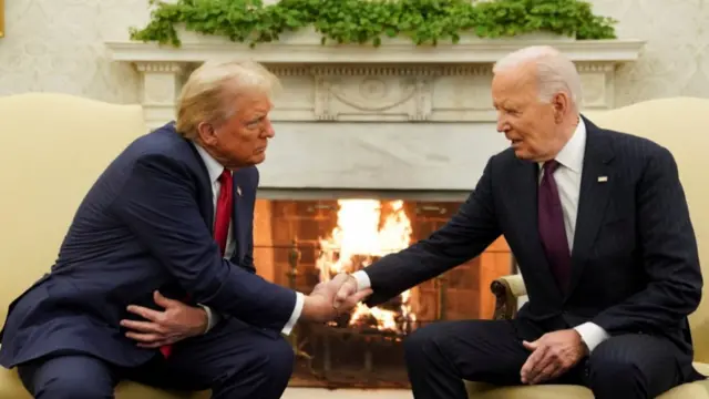 Donald Trump (L) and Joe Biden (R) shake hands during meeting inside Oval Office. Both are sitting down on cream-coloured chairs in front of the fireplace. Trump is in a dark blue suit with white shirt and red tie, while Biden is in a black pin-striped suit with white shirt and purple tie