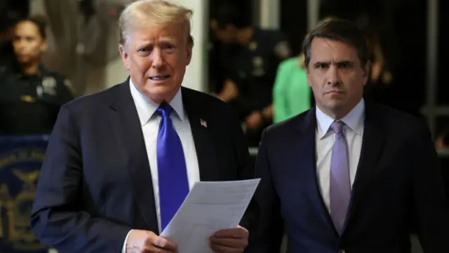 Donald Trump, in a suit and tie, holds papers and stands beside his attorney Todd Blanche, also in a suit and tie, in May 2024