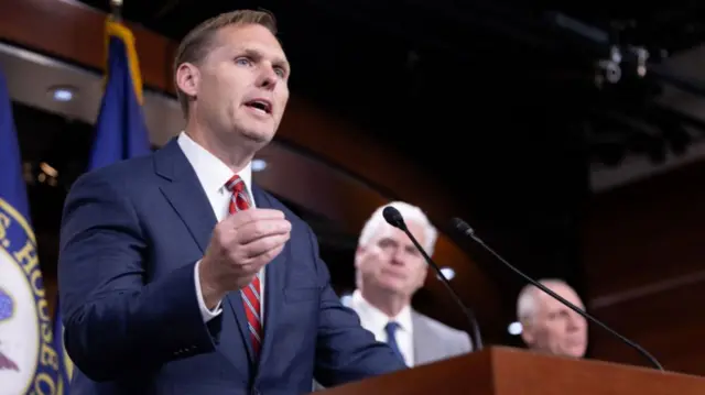 Republican Congressman Michael Guest speaks into a microphone and gestures with his hand - he's wearing a blue suit with a rep and white striped tie