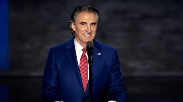 Man stands at podium with slicked back grey hair wearing a navy suit and red tie