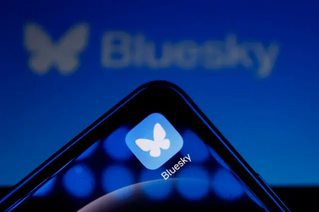 Bluesky logo in background with a handheld device showing the app logo of a white butterfly on a blue square