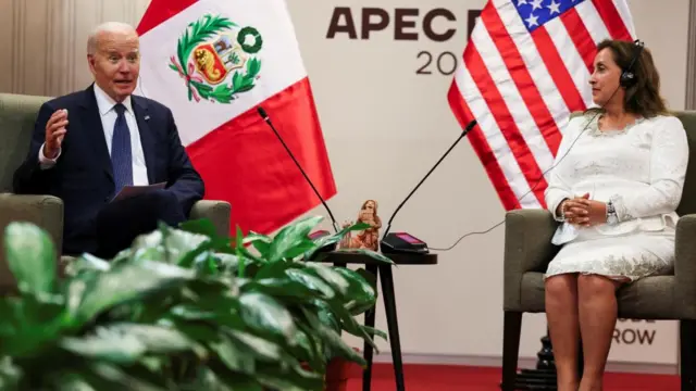 Biden sits in an armchair on the left, gesturing as he speaks. Boluarte sits on the right, wearing headphones and watching him. Behind them the US and Peruvian flags are on display