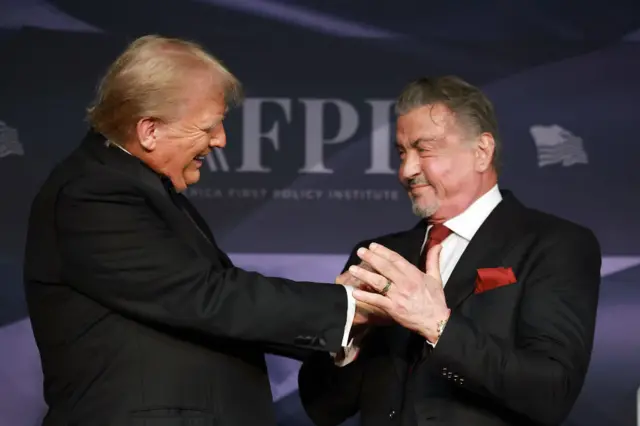 Two men smile at each other while wearing tuxedos and grasping each others hands