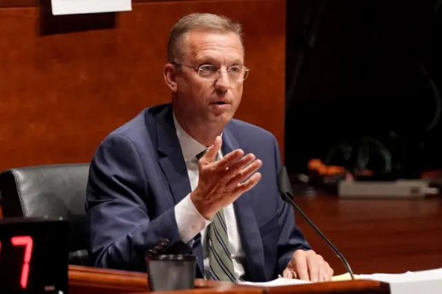 Man in blue suit and silver ties gestures his right hand while sitting and speaking at a microphone