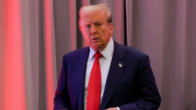 Donald Trump mid-shot cutting off below the waist. He's in a dark blue suit, white shirt and red tie. Behind him is a set of heavy dark-grey stage curtains illuminated by a red light to the left