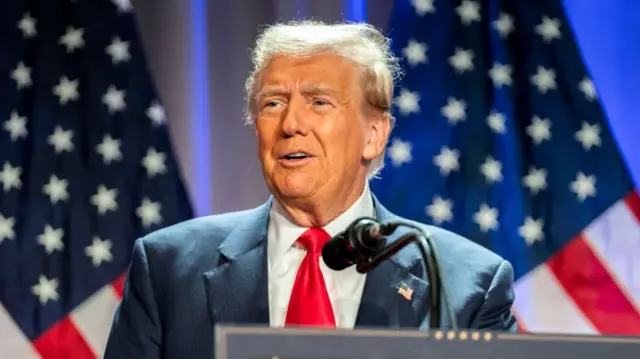 Center shot of Donald Trump on stage, cut off below the shoulders. He wears a dark blue suit, white shirt and red tie, with an American flag pin on the left label. Two faint American flags can be seen in the background