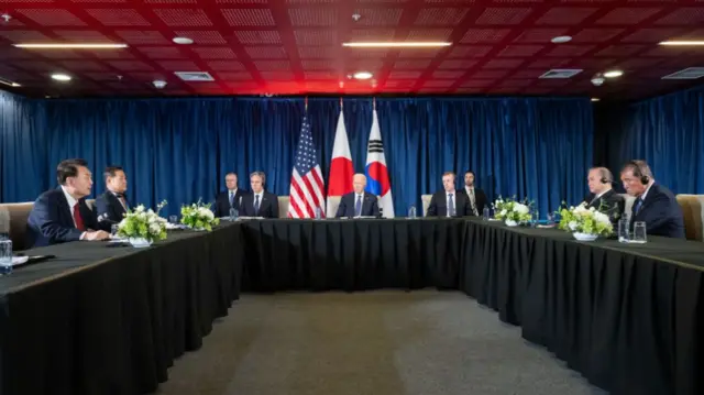 Joe Biden during bilateral summit with Yoon Suk Yeol (L) and Shigery Ishiba (R). They're all sitting around a table covered with black tablecloth. Aides and translators also at the table