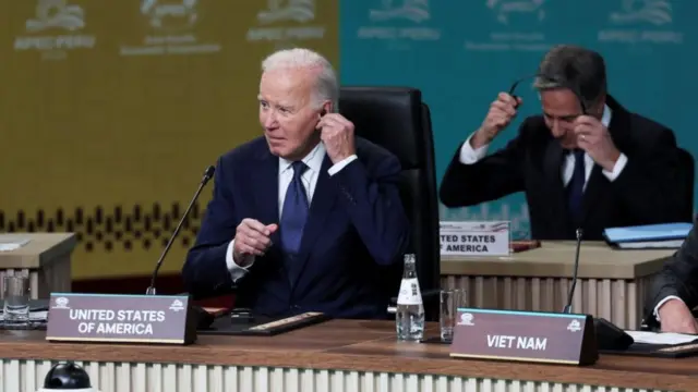 Joe Biden puts headphone in as he prepares for the first Apec session. He's sitting down in a dark blue suit, white shirt and blue tie. Behind him, to his left, is Antony Blinken putting on his own headphones