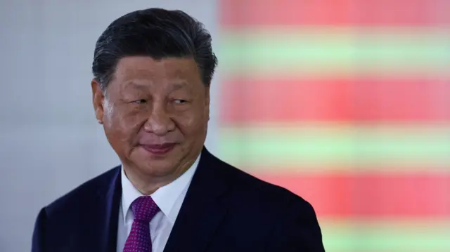 Xi Jinping smirking in dark suit, whit shirt and purple patterned suit. His head is tilted to the left, background is blurred