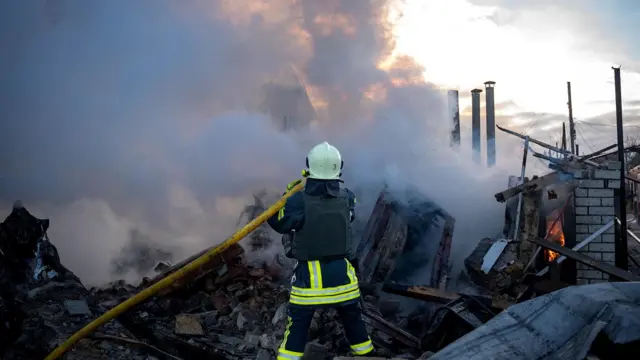 Firefighter stands with a hose over rubble in front of a fire and smoke