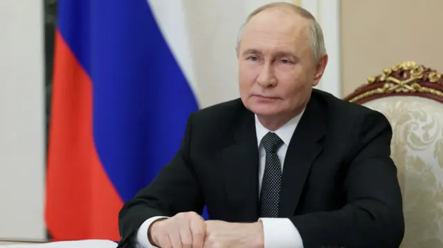 Vladimir Putin in a suit and tie sat at a desk. Behind him is Russia's flag.