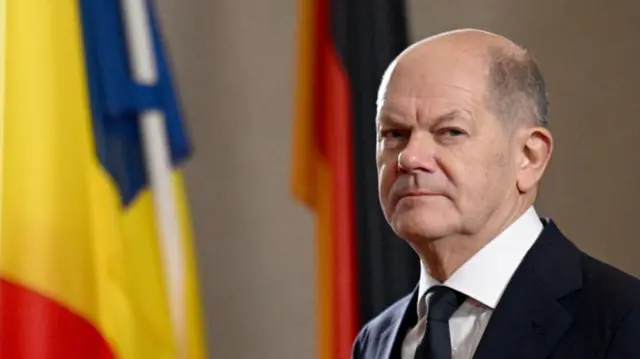 German chancellor Olaf Scholz to the right of the frame, mid shot cutting off below shoulders. He's in a black suit with white shirt and black tie. In the background, blurred, are a German flag and a Romanian one