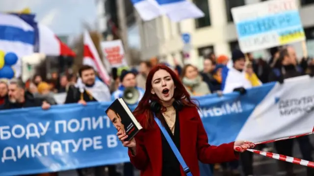 A woman stands in front of a Russian banner, holding a copy of Alexei Navalny's book and calling out. A large crowd is behind her with blue and white flags and