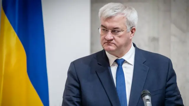 Ukraine's foreign affairs minister Andrii Sybiha at a press conference. Shot cuts off mid-chest, he's wearing a dark grey suit with white shirt and blue tie as he looks to his right. To the left of the image is a Ukrainian flag