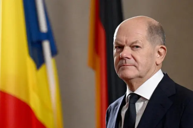 Close up shot of German chancellor Olaf Scholz, there are some flags behind him