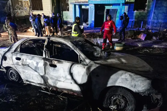 A burnt out car in the region of Sumy, Ukraine. The area was struck by Russian missiles and people behind the car inspect the damage to buildings nearby