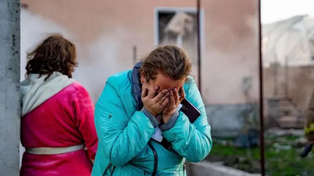 A woman cries into her hands, standing in front of a damaged building